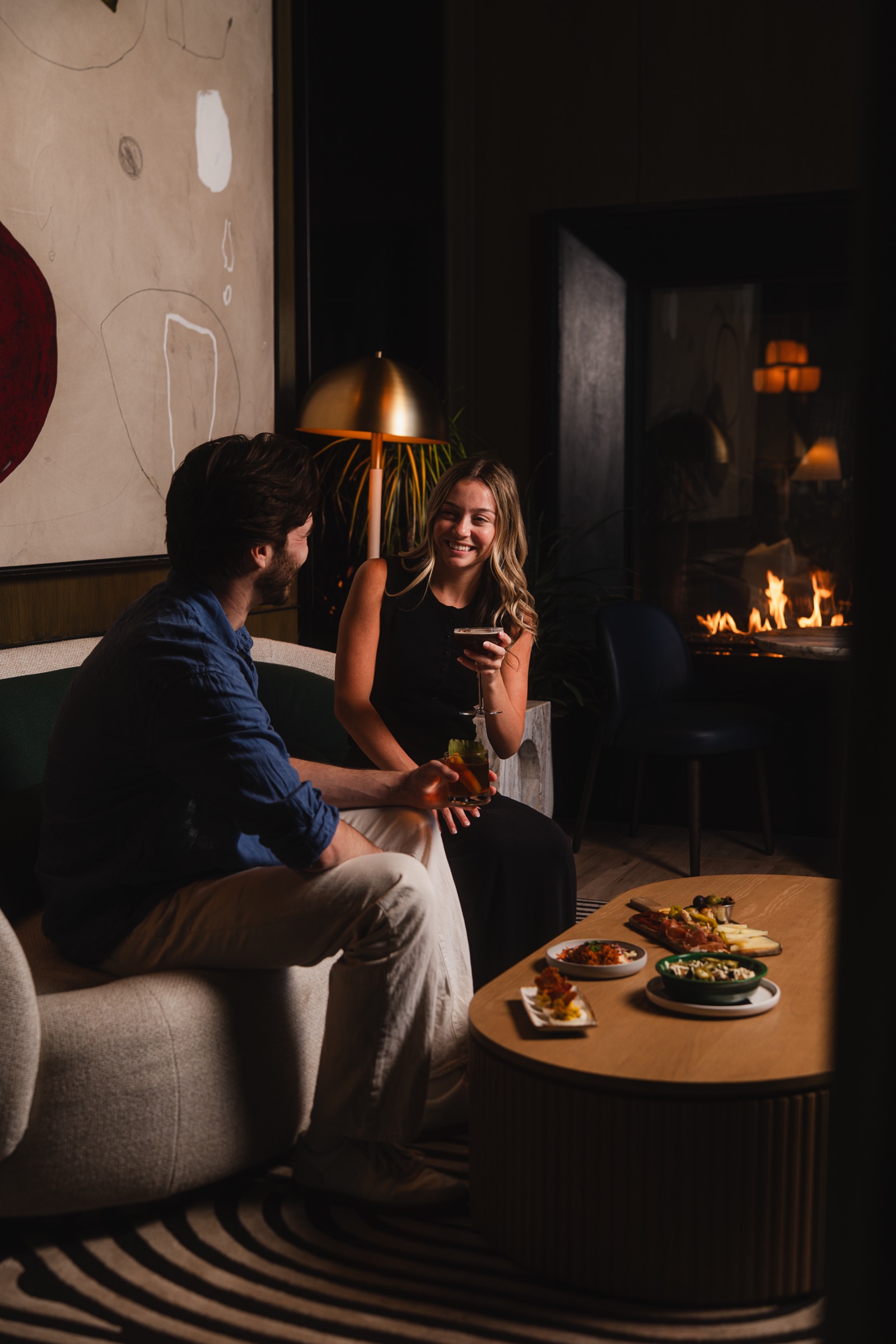 Couple enjoying cocktails and plates beside a fireplace in an intimate lounge