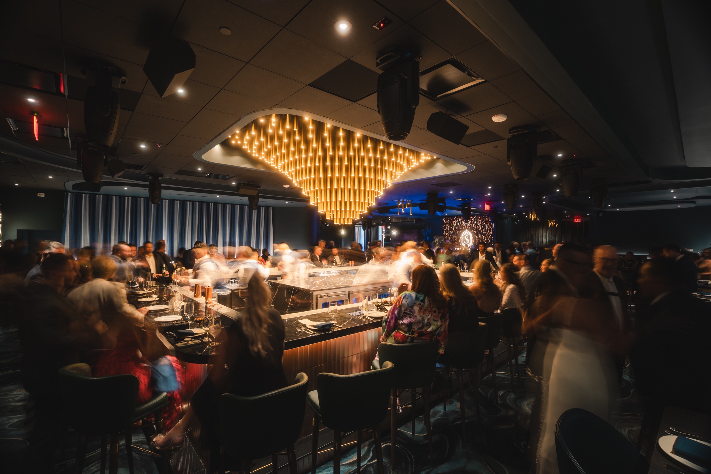 Busy cocktail bar captured in motion beneath a gold light installation
