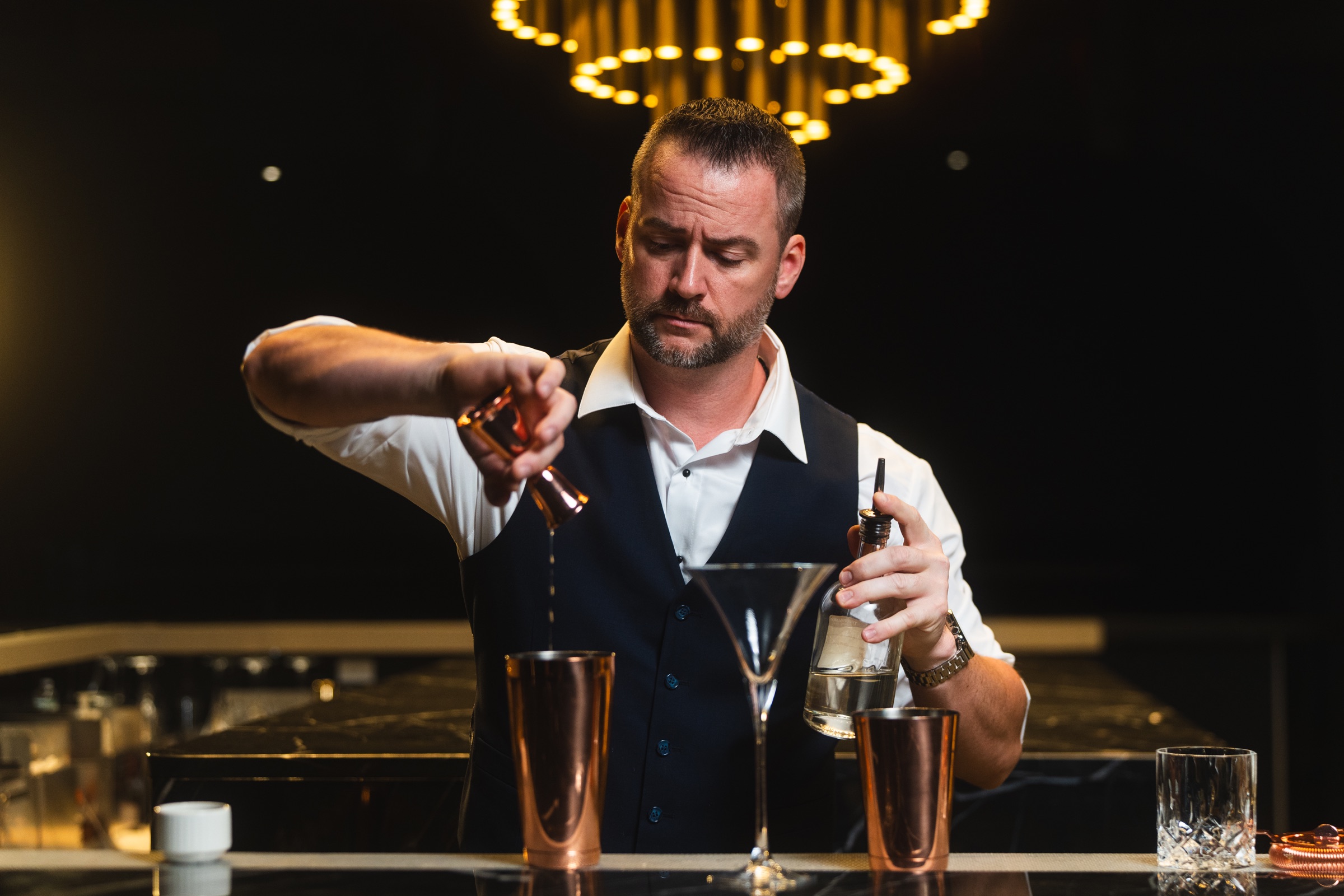 Bartender measuring ingredients beneath sculptural pendant lights